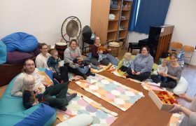 Eltern und kleine Kinder sitzen gemeinsam auf bunten Matten in einem Förderraum, umgeben von Musikinstrumenten. Die Szene zeigt eine inklusive Eltern-Kind-Gruppe der Frühförderung für blinde und sehbehinderte Kinder.