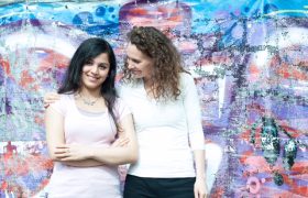 Eine Jugendliche und eine Mentorin stehen vor einer bunten Wandmalerei; die Mentorin legt unterstützend den Arm um die Jugendliche. Das Foto steht für die Bildungs- und Mentoringprogramme von SchlauFox für benachteiligte Kinder und Jugendliche