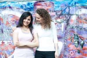 Eine Jugendliche und eine Mentorin stehen vor einer bunten Wandmalerei; die Mentorin legt unterstützend den Arm um die Jugendliche. Das Foto steht für die Bildungs- und Mentoringprogramme von SchlauFox für benachteiligte Kinder und Jugendliche